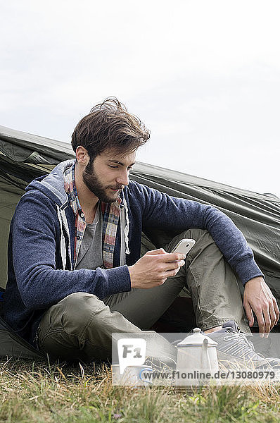 Mann benutzt Smartphone vor dem Zelt auf einem Hügel vor klarem Himmel