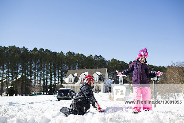 Siblings playing with snow on field against clear blue sky