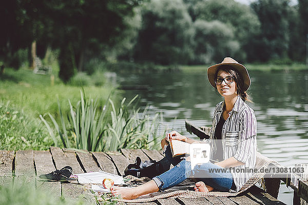 Porträt einer Frau mit Buch  die auf einem Steg vor einem See im Wald sitzt