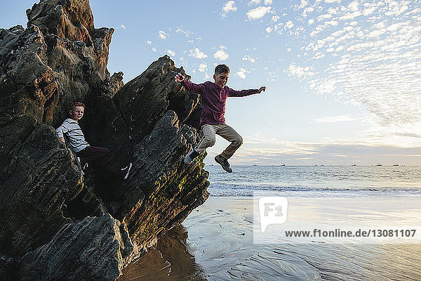 Playful boy jumping at beach while brother sitting on rocks