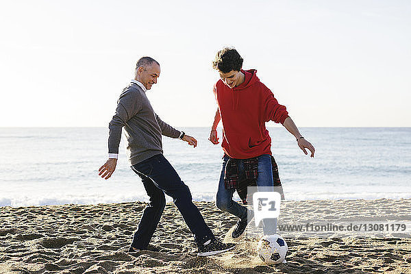 Happy father and son playing soccer at beach against clear sky