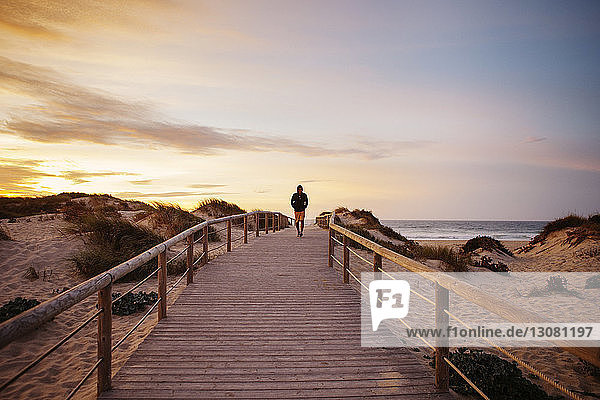 Distant view of hiker walking on footbridge at beach against sky during sunset