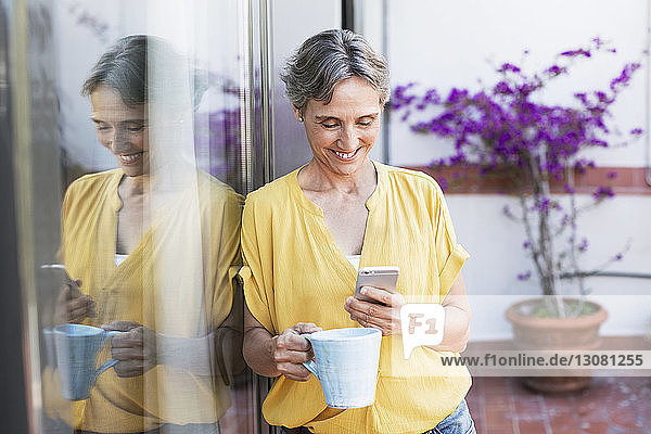 Glückliche reife Frau benutzt Smartphone  während sie eine Kaffeetasse auf der Veranda hält