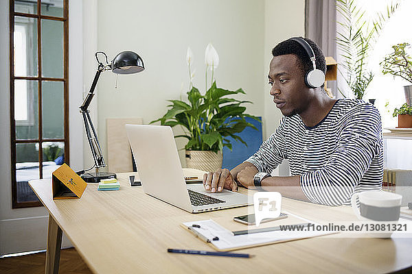 Junger Mann hört Musik  während er den Laptop im Heimbüro benutzt