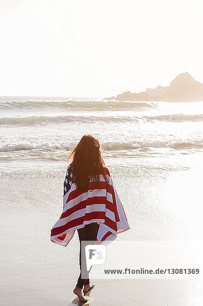 Rückansicht einer in eine amerikanische Flagge gehüllten Frau  die am Strand am Ufer spazieren geht