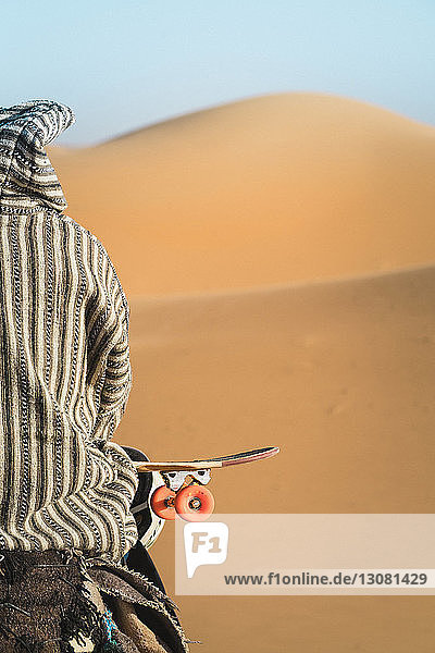 Rückansicht eines Mannes mit Skateboard  der bei Sonnenschein auf einem Kamel sitzt