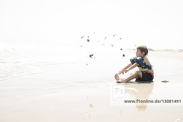 Side view of boy playing with sand at beach against sky during sunny day
