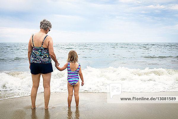 Rear view of granddaughter and grandmother standing on shore at beach