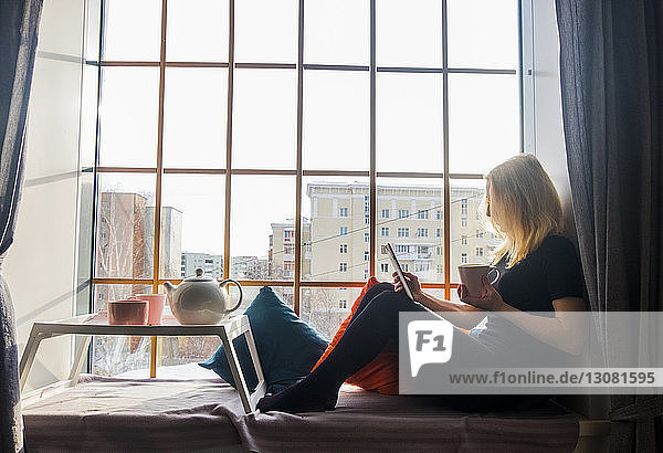 Frau benutzt Tablet-Computer  während sie zu Hause am Fensterplatz in einer Nische trinkt
