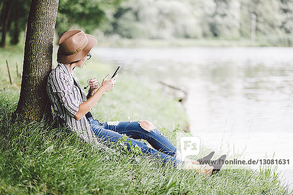 Frau benutzt Mobiltelefon  während sie auf einem Grasfeld am Seeufer im Wald sitzt