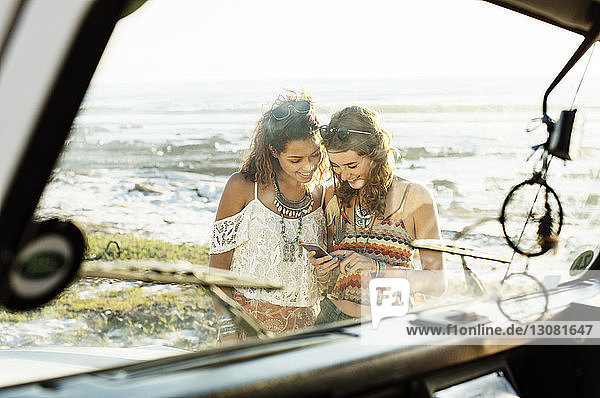 Happy friends using mobile phone at beach seen through windshield