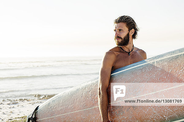 Mann ohne Hemd mit Surfbrett am Strand bei klarem Himmel am sonnigen Tag