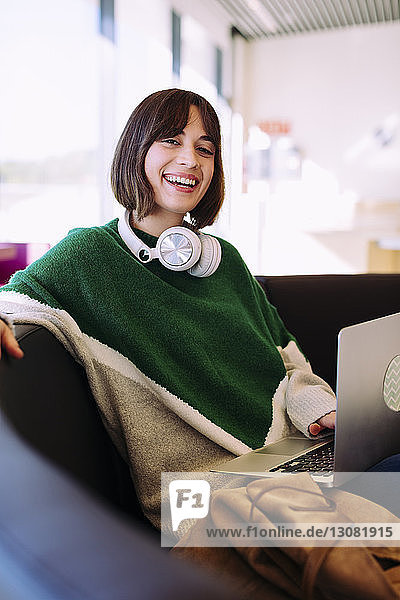Portrait of smiling woman using laptop computer while sitting on sofa in library