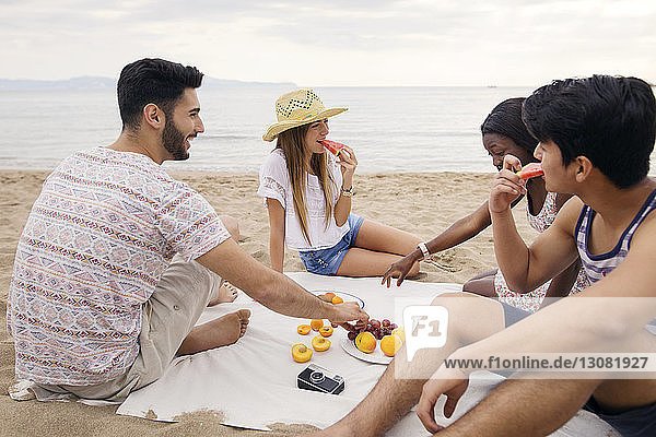 Happy friends eating fruits while sitting on beach