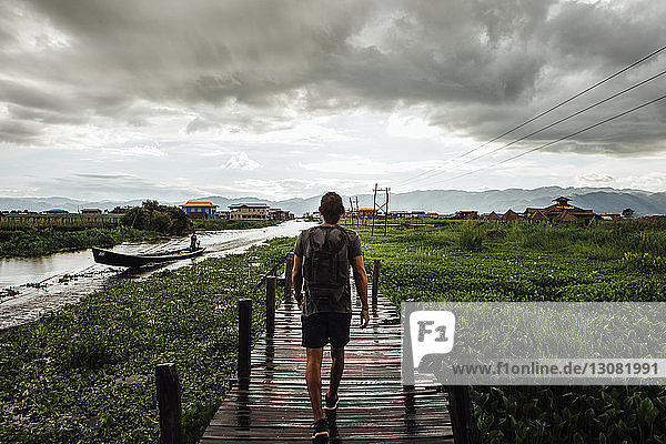Rear view of man with backpack walking on footbridge by Inle lake against cloudy sky