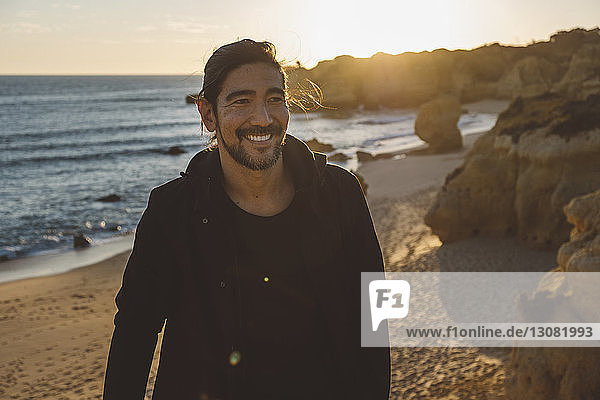 Smiling man looking away while standing at beach against sky during sunset
