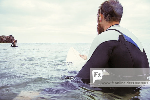 Rückansicht eines männlichen Surfers  der auf einem Surfbrett im Meer vor bewölktem Himmel sitzt