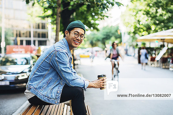 Porträt eines glücklichen Mannes  der einen Einwegbecher hält  während er auf einer Bank auf dem Bürgersteig sitzt