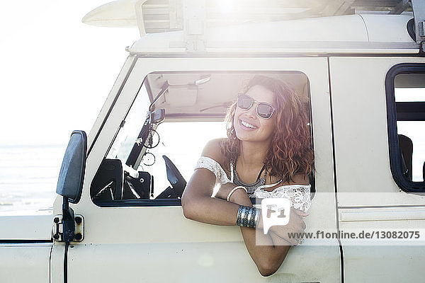 Happy woman in sunglasses looking through car window during sunny day
