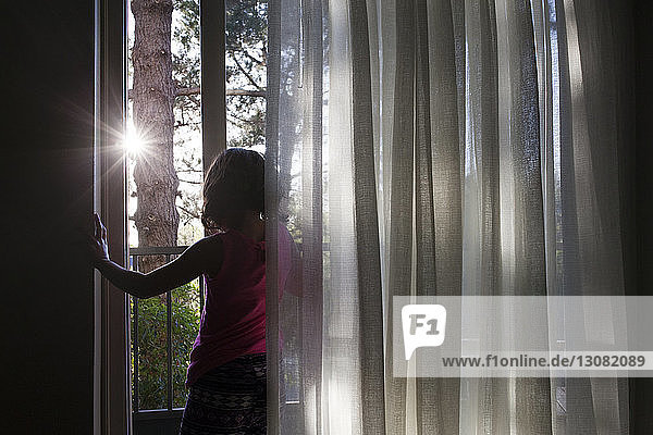 Rückansicht eines Mädchens  das an einem sonnigen Tag am Fenster bei Vorhängen steht
