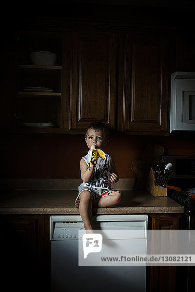 Porträt eines Jungen  der Banane isst  während er zu Hause auf dem Küchentisch sitzt