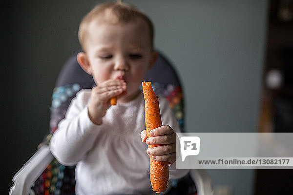 Süßes Mädchen isst Karotte  während es zu Hause auf einem Hochstuhl sitzt
