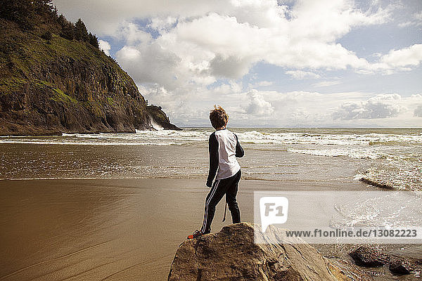Rear view of boy standing on rock at seashore against cloudy sky