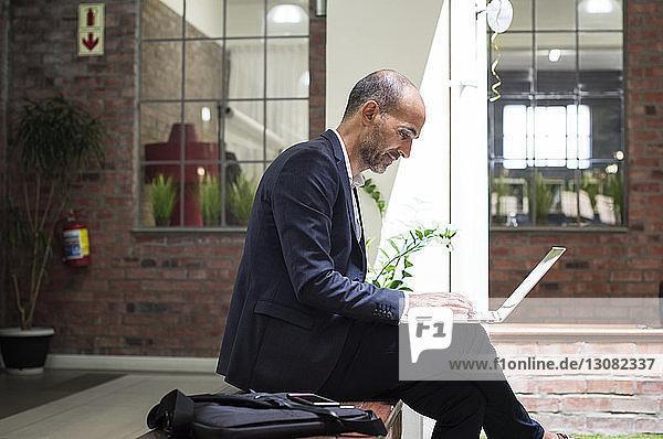 Side view of businessman using laptop computer while sitting on seat against office