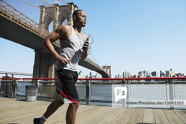 Man listening music and jogging on boardwalk against Brooklyn Bridge