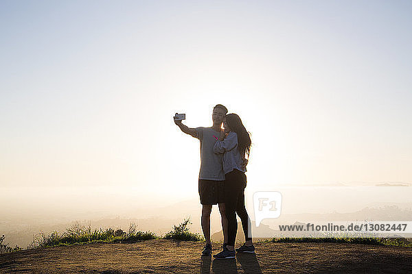 Couple taking selfie through smart phone while standing on mountain against clear sky