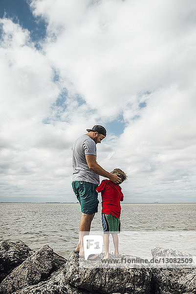 Vater mit Sohn steht auf Felsen am Strand vor bewölktem Himmel