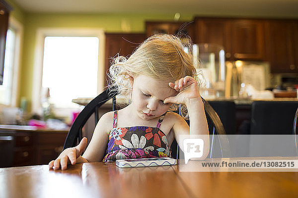 Mädchen betrachtet Mobiltelefon  während sie zu Hause auf einem Stuhl am Tisch sitzt