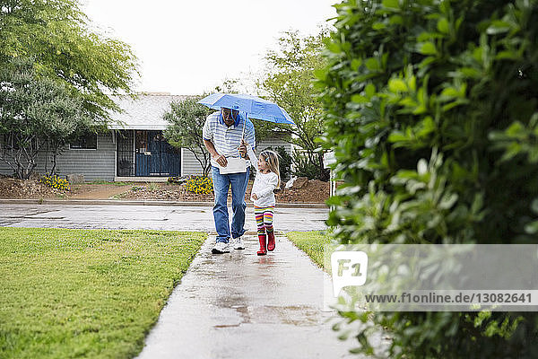 Grandfather and granddaughter walking under umbrella on footpath at park