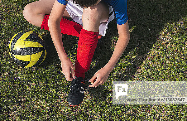 Hochwinkelaufnahme eines Jungen beim Schnürsenkelbinden  der neben einem Fussball auf einem Rasenplatz sitzt