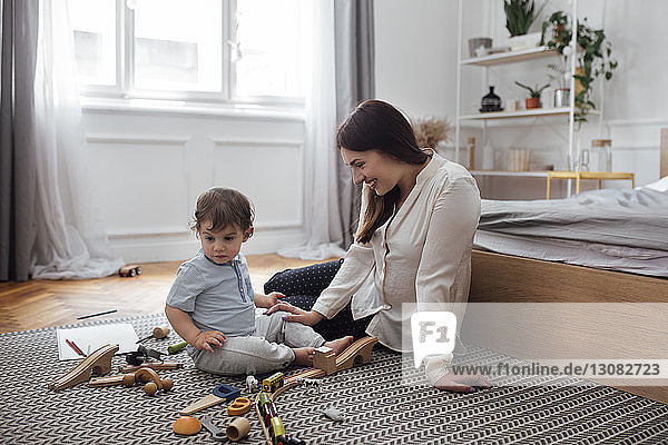 Happy mother looking at baby boy playing with toys on rug in bedroom