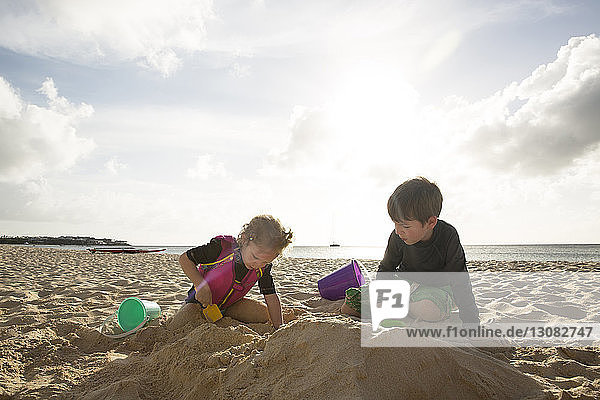 Siblings playing with sand at beach against sky