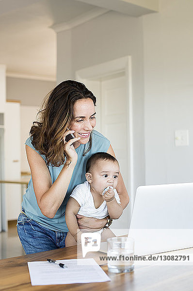 Mother talking on phone while carrying baby boy looking at laptop computer