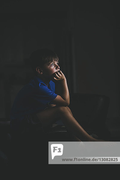 Seitenansicht eines nachdenklichen Jungen mit Hand am Kinn  der auf einem Stuhl in der Dunkelkammer sitzt