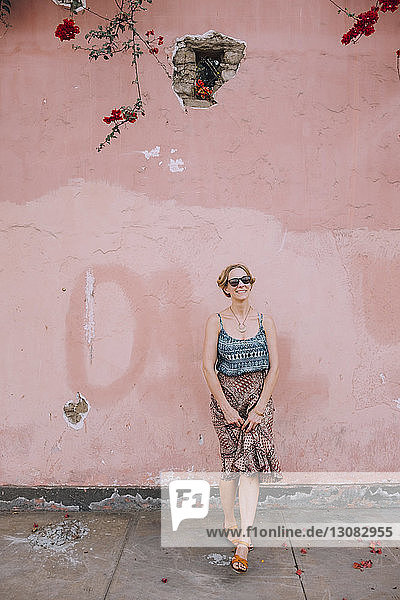 Porträt einer lächelnden Frau mit Sonnenbrille  die auf dem Fußweg an der Wand steht