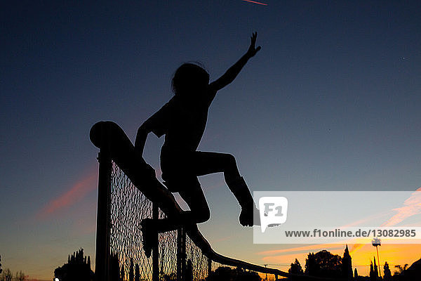 Silhouette eines sportlichen Mädchens  das in der Abenddämmerung gegen den Himmel über den Zaun springt