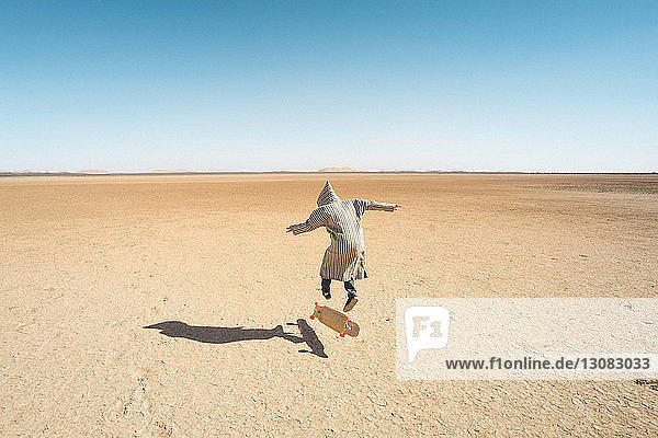 Rückansicht eines Mannes beim Skateboarden auf Sand vor klarem blauen Himmel