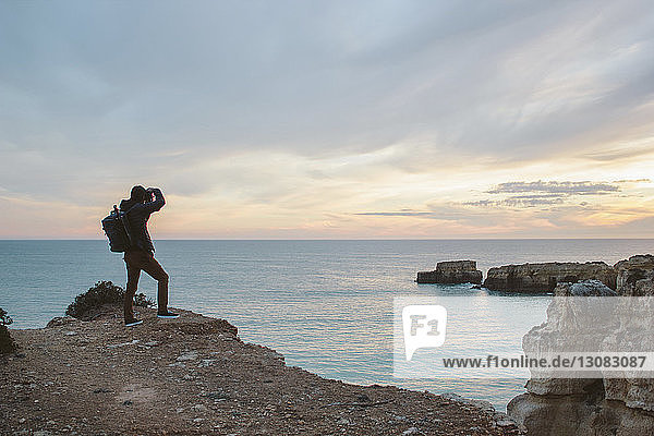 Man photographing sea while standing on rock formation against cloudy sky during sunset