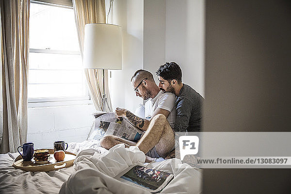 Gay men reading newspaper while sitting on bed at home