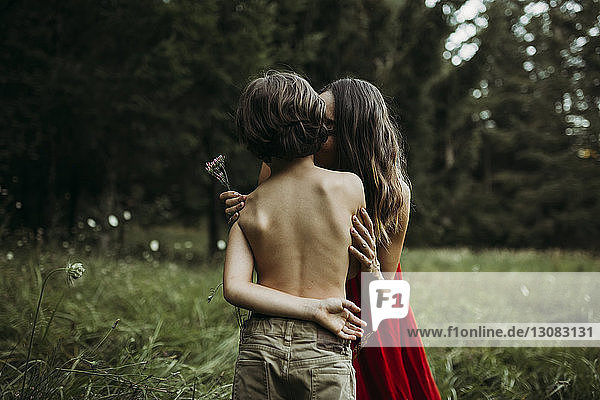 Mother kissing shirtless son while holding flowers at grassy field
