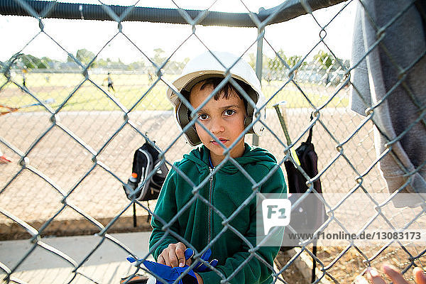 Portrait of boy wearing baseball helmet while sitting in dugout seen through chainlink fence