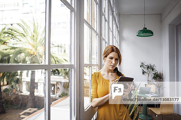 Businesswoman using tablet computer while leaning on window in office