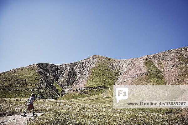 Boy walking on trail in field by mountains against clear blue sky