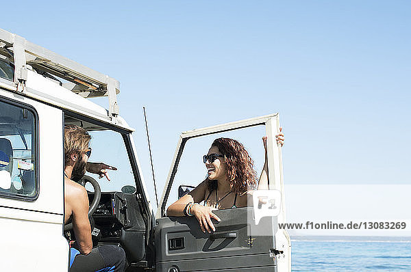 Frau unterhält sich mit einem männlichen Freund  der an einem sonnigen Tag im Geländewagen am Strand sitzt