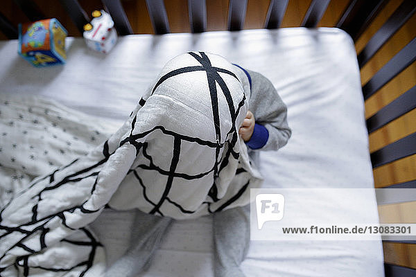 Overhead view of playful baby boy hiding in blanket while playing peekaboo in crib at home