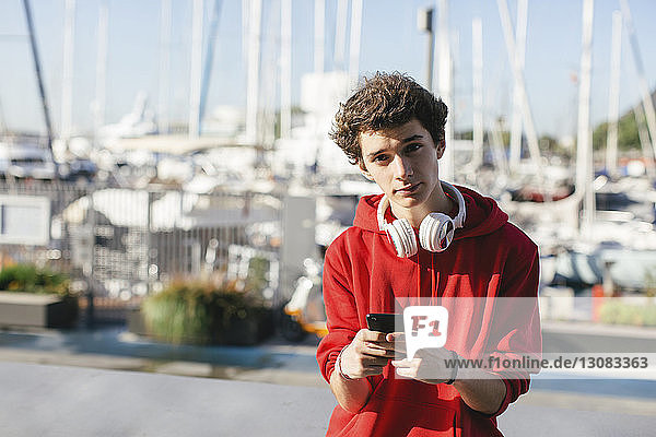 Portrait of confident teenage boy holding smart phone against boats during sunny day